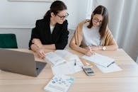 Two women at a desk studying with books, notes, and a laptop, emphasizing collaboration.